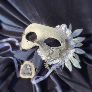 Puede incluir: Máscara blanca de carnaval con detalles intrincados. La máscara presenta una superficie texturizada, adornada con encaje, engranajes metálicos y detalles de cristal. Hojas y cintas plateadas completan el diseño. Sobre un fondo de terciopelo azul oscuro.
