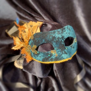 Puede incluir: Una máscara de terciopelo azul verdoso con detalles dorados y un diseño de ojo de cerradura. La máscara está decorada con engranajes dorados y una pequeña pluma.