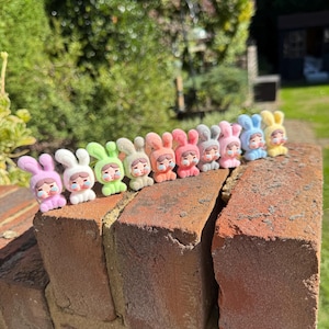 May include: A collection of small bunny figurines in various pastel colours, including pink, green, and blue. Each bunny has a sad expression and wears a different outfit. They are arranged on a weathered brick wall, with a blurred green backdrop.