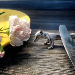 Peut inclure: Une figurine de blaireau en argent et un couteau à beurre en argent avec une couronne et le texte "U.B. Germany" sur le manche. Le couteau est posé sur une surface en bois à côté de la figurine. Une assiette jaune avec des œillets blancs est en arrière-plan.