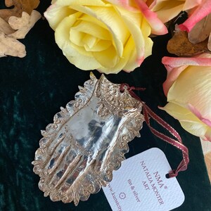 Könnte beinhalten: Silberfarbener, handförmiger Schmuck mit filigranen Details, aufgehängt an einem weinroten Band. Der Schmuck liegt auf einem dunkelgrünen Samtgrund, mit gelben und rosa Rosen und getrockneten Blättern im Hintergrund. Ein Etikett mit "NATALIA MONTER ART".