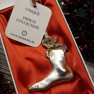 Könnte beinhalten: Silberfarbener Weihnachtsstrumpf-Ornament in einer weißen Schachtel mit rotem Satinfutter. Das Ornament hat ein goldenes Band zum Aufhängen. Das Etikett lautet "UNIQUE DESIGN COLLECTION" und die Website-Adresse www.kunstcity.de.