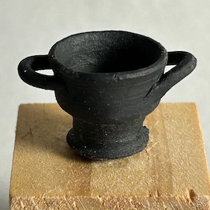 May include: A small, black, ceramic cup with two handles. The cup has a textured surface and sits on a wooden block. The cup's design is reminiscent of ancient pottery.