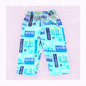 May include: Light blue fleece pants with a Hanukkah theme. The pants feature menorahs, stars of David, and the words "Light," "Oh Joy," and "Love." The pants have a comfortable elastic waistband.