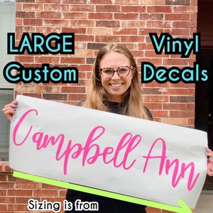 May include: A white vinyl decal with the name "Campbell Ann" written in pink cursive lettering. The decal is 30 inches wide and is shown being held by a person. The text "Sizing is from LEFT to RIGHT 30 inch shown here" is written below the decal.