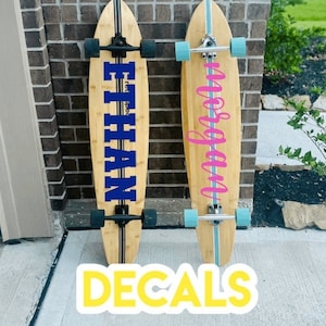 Könnte beinhalten: Zwei Holz-Skateboards mit individuellen Aufklebern. Ein Skateboard hat einen blauen Aufkleber mit dem Namen "ETHAN" und das andere einen rosa Aufkleber mit dem Wort "Morgan".