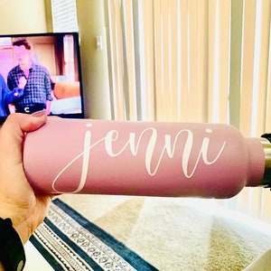 K&ouml;nnte beinhalten: Eine rosa Wasserflasche mit einem wei&szlig;en Vinyl-Aufkleber, auf dem "jenni" steht.