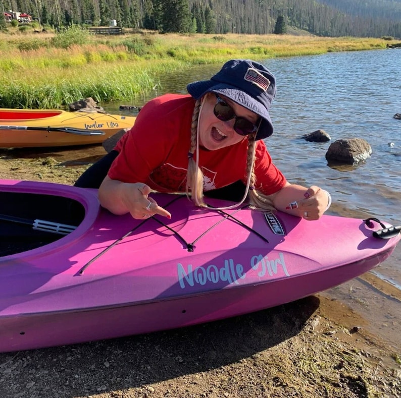 May include: A woman wearing a red shirt and a blue hat with an American flag patch is sitting in a pink kayak. The kayak has the words "Noodle Girl" written on the side.