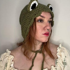 May include: A green crochet frog hat with white and black ear detailing. The hat features a tie closure. This whimsical hat is a fun accessory.