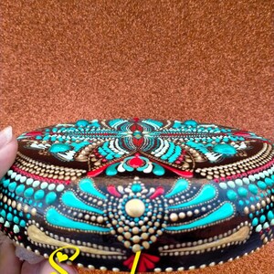 Op de afbeelding: Een ronde, bruine steen beschilderd met een kleurrijk mandala-ontwerp. De mandala heeft turquoise, gouden, rode en witte stippen en swirls.