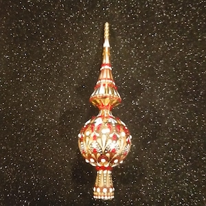 May include: A gold and red Christmas tree topper with a detailed design. The topper has a conical shape with a pointed top and a round base. The design includes red and white accents.