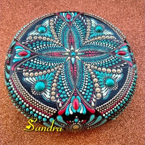 Op de afbeelding: Een ronde, zwarte steen beschilderd met een mandala-ontwerp. De mandala heeft turquoise, witte, gouden en rode stippen en swirls. De steen is gesigneerd met "Sandra".