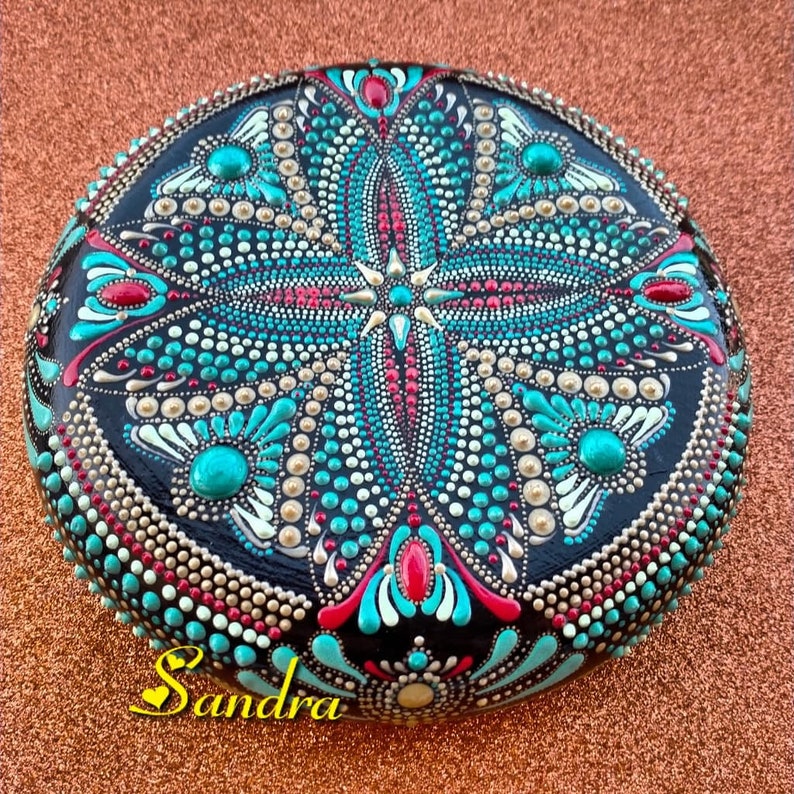 Op de afbeelding: Een ronde, zwarte steen beschilderd met een mandala-ontwerp. De mandala heeft turquoise, witte, gouden en rode stippen en swirls. De steen is gesigneerd met "Sandra".