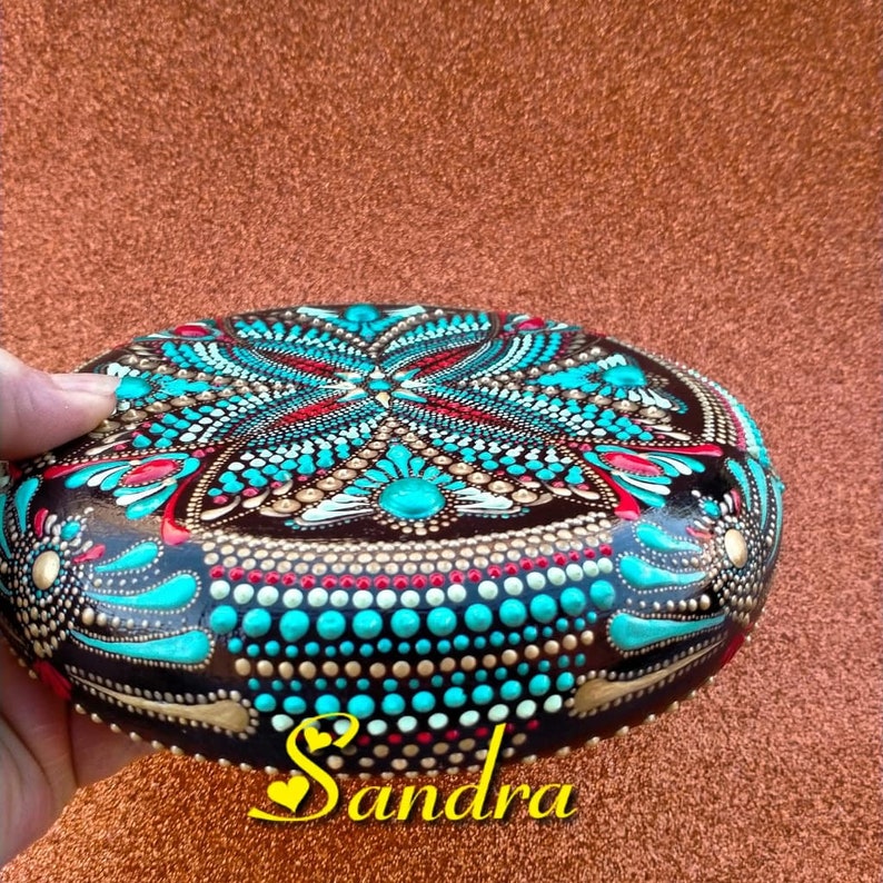 Op de afbeelding: Een ronde, bruine steen beschilderd met een mandala-ontwerp in turquoise, goud, wit en rood. De steen is versierd met stippen en heeft een glanzende afwerking. Het woord "Sandra" staat in geel op de onderkant van de steen.