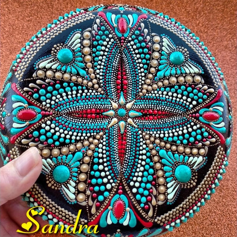 Op de afbeelding: Een ronde, zwarte steen beschilderd met een mandala-ontwerp. De mandala heeft een kleurenschema in turquoise, rood, wit en goud. De steen is versierd met stippen en swirls.