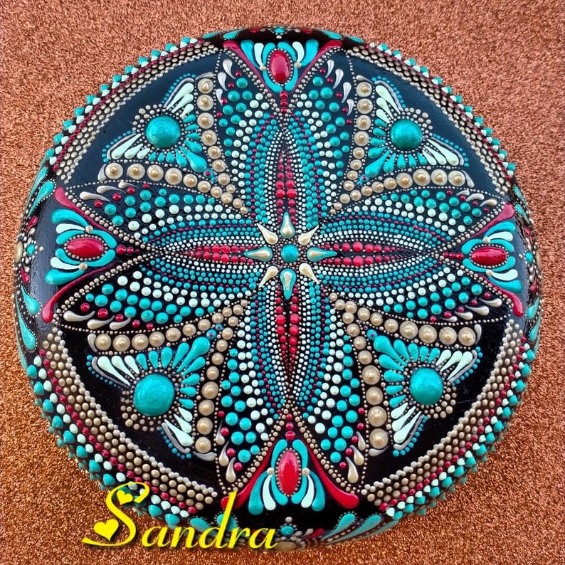 Op de afbeelding: Een ronde, zwarte steen beschilderd met een mandala-ontwerp. De mandala heeft een turquoise, rood, goud en wit kleurenschema. De steen is versierd met stippen en swirls.