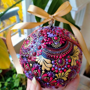 May include: A hand-painted, round ornament with a black base, adorned with intricate patterns in shades of pink, purple, and gold. The ornament is topped with a gold ribbon for hanging. The design features a dot-based mandala style.