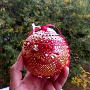 May include: A red Christmas ornament with gold and white decorative accents. The ornament has a textured surface with a pattern of dots, swirls, and floral designs. A red ribbon is attached for hanging.