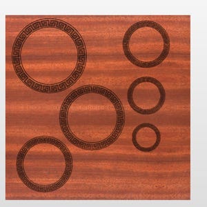 Puede incluir: Una superficie de madera de color marrón rojizo con un patrón de vetas. Seis diseños circulares de color marrón oscuro con un borde de patrón de clave griega están dispuestos en la superficie. Los círculos varían en tamaño.
