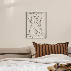 May include: A white quilted bedspread with a brown and tan striped throw pillow and a brown throw blanket on a bed with a black and white line design throw pillow. A black floor lamp with a black shade is in the background. There is a stack of books on a wooden table in the foreground.