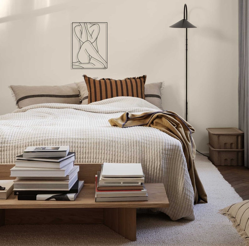 May include: A white quilted bedspread with a brown and tan striped throw pillow and a brown throw blanket on a bed with a black and white line design throw pillow. A black floor lamp with a black shade is in the background. There is a stack of books on a wooden table in the foreground.