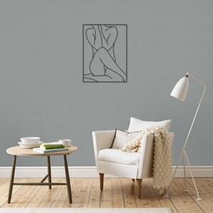May include: A black metal wall sculpture depicting a line drawing of a person sitting with their legs crossed. The sculpture is hanging on a gray wall in a living room setting.