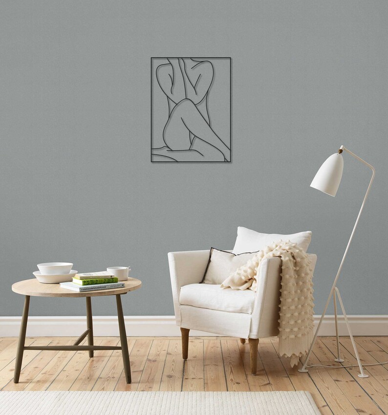 May include: A black metal wall sculpture depicting a line drawing of a person sitting with their legs crossed. The sculpture is hanging on a gray wall in a living room setting.