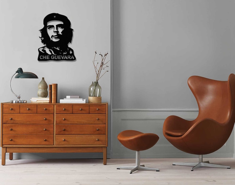 May include: A black and white silhouette of Che Guevara's face with the text "CHE GUEVARA" below. The image is displayed on a wall above a wooden dresser with drawers. A brown leather egg chair and a matching ottoman are in the foreground.
