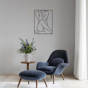 May include: A black line art wall decor featuring a woman sitting with her arms around her legs. The decor is hanging on a light gray wall above a blue armchair and ottoman.