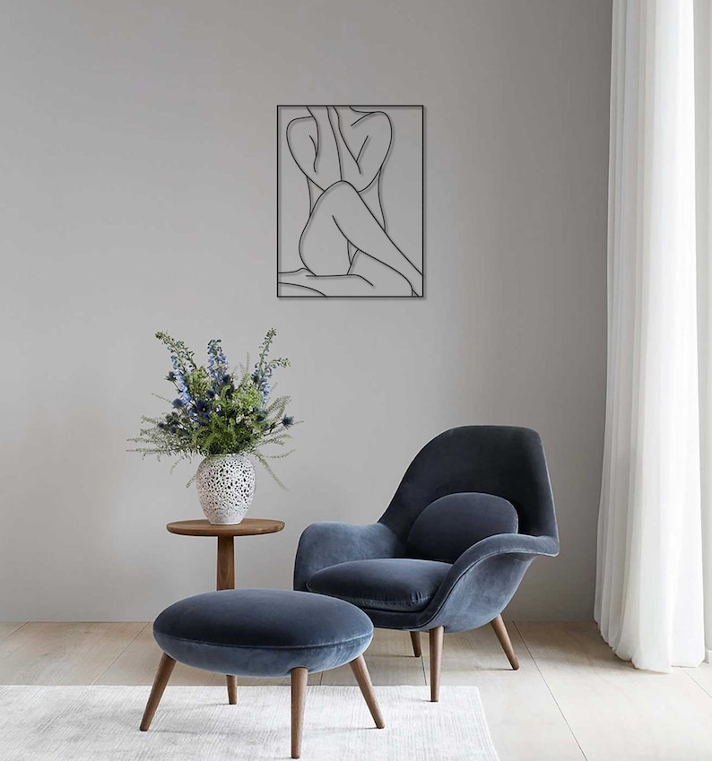 May include: A black line art wall decor featuring a woman sitting with her arms around her legs. The decor is hanging on a light gray wall above a blue armchair and ottoman.