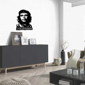May include: Black and white silhouette wall art of Che Guevara, a famous revolutionary, with the text "CHE GUEVARA" below the image. The art is displayed on a white wall above a dark grey cabinet with wooden legs.