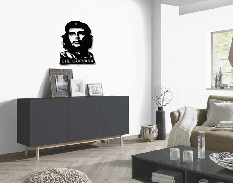 May include: Black and white silhouette wall art of Che Guevara, a famous revolutionary, with the text "CHE GUEVARA" below the image. The art is displayed on a white wall above a dark grey cabinet with wooden legs.