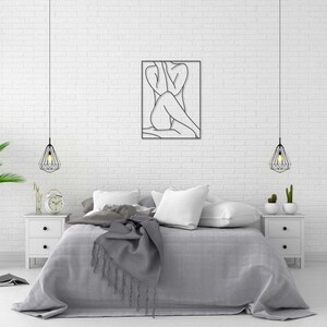 May include: A bedroom interior with a white brick wall, a bed with gray bedding, a black metal line art wall hanging of a person, two hanging pendant lights, and two white nightstands with lamps.