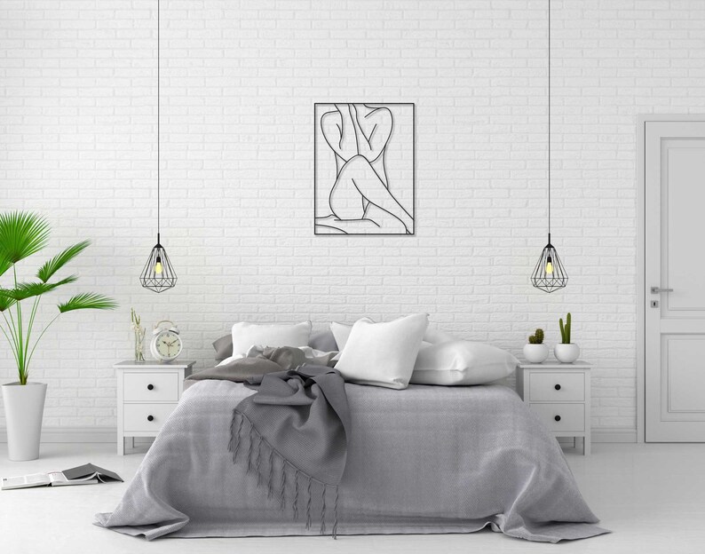 May include: A bedroom interior with a white brick wall, a bed with gray bedding, a black metal line art wall hanging of a person, two hanging pendant lights, and two white nightstands with lamps.