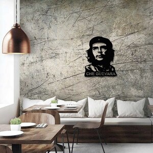 May include: Black and white silhouette of Che Guevara's face with the text "CHE GUEVARA" below. The silhouette is mounted on a gray textured wall in a dining room setting with a wooden table and chairs.