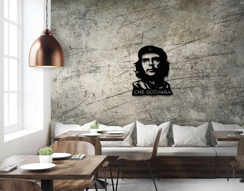 May include: Black and white silhouette of Che Guevara's face with the text "CHE GUEVARA" below. The silhouette is mounted on a gray textured wall in a dining room setting with a wooden table and chairs.