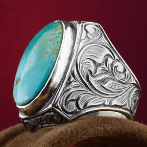 May include: A silver ring with a turquoise stone set in a bezel setting. The ring band is intricately engraved with a floral design.