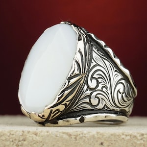 May include: A silver ring with a white oval stone and intricate floral engravings. The ring is set against a red background.