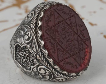 May include: A silver ring with an ornate design and a red stone set in the center. The stone features an intricate design of a six-pointed star with Arabic script.