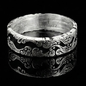 May include: A silver ring with a detailed wave pattern. The ring has a dark, oxidized background that highlights the intricate design. The ring's surface reflects light, creating a contrast between the bright silver and the dark recesses of the wave design.