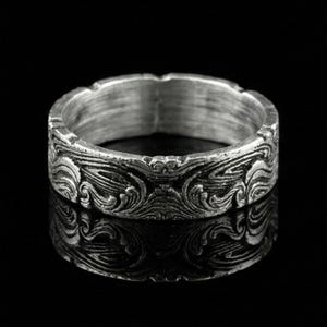 May include: A silver ring with an ornate, swirling design. The ring showcases a detailed pattern of flowing lines and curves, giving it a textured look. The ring is set against a black backdrop, accentuating its metallic shine.