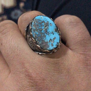 May include: A silver ring with a large, oval turquoise stone. The stone has a mottled appearance with shades of blue and brown. The ring has a gold-colored border around the stone and ornate silver detailing on the band.