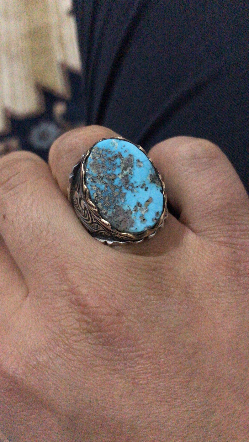 May include: A silver ring with a large, oval turquoise stone. The stone has a mottled appearance with shades of blue and brown. The ring has a gold-colored border around the stone and ornate silver detailing on the band.