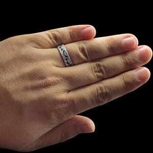 May include: A silver ring with an intricate, swirling pattern. The ring is worn on a hand against a black background. The design appears to be etched or engraved, creating a detailed and textured surface. The ring's band is approximately 0.25 inches wide.