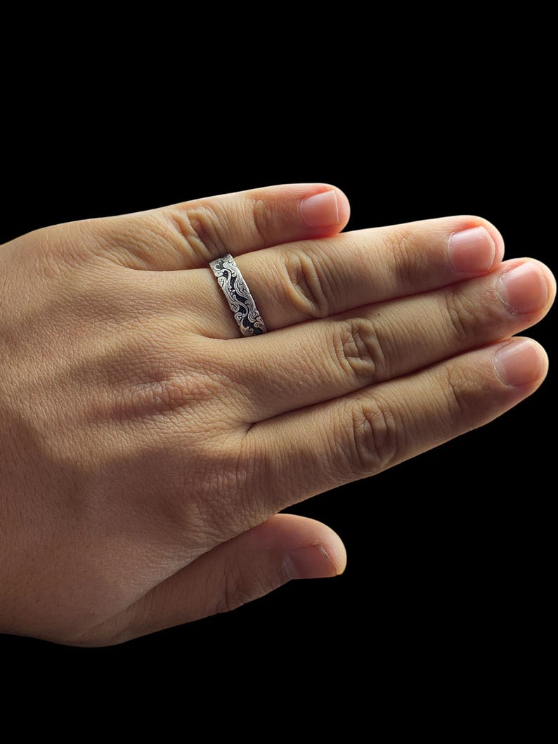 May include: A silver ring with an intricate, swirling pattern. The ring is worn on a hand against a black background. The design appears to be etched or engraved, creating a detailed and textured surface. The ring's band is approximately 0.25 inches wide.