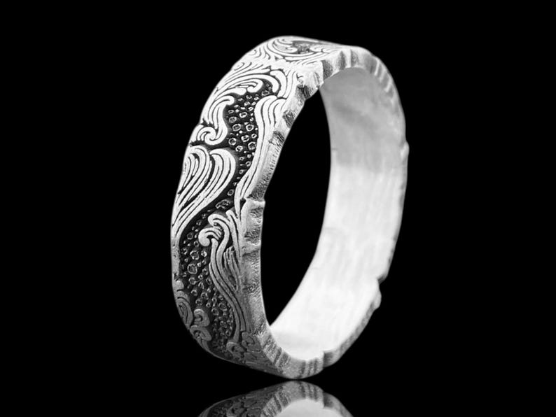 May include: A silver ring with an ornate, engraved floral pattern. The ring has a textured, uneven outer edge and a polished inner surface. The design features swirling lines and small, circular details, set against a dark background.