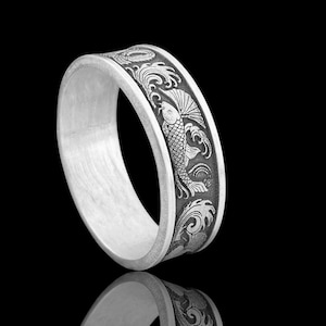 May include: A silver ring with a detailed engraved design of a koi fish swimming through waves. The ring has a wide band and a polished finish, reflecting light. The design is dark against the silver, creating a contrast.