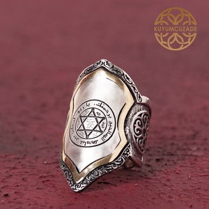 May include: Silver ring with a shield-shaped design featuring a six-pointed star and Arabic script. The ring has a gold trim and intricate details.
