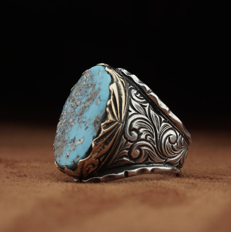 May include: A silver ring with a large, oval turquoise stone. The stone has a mottled appearance with shades of blue, gray, and brown. The ring's band features intricate, engraved floral patterns and gold accents, creating a vintage aesthetic.