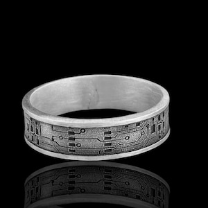 Könnte beinhalten: Ein silberner Ring mit einem Leiterplatten-Design. Der Ring hat ein flaches Band mit geätzten Linien und geometrischen Formen, was eine technikinspirierte Ästhetik erzeugt. Der Ring wird vor einem schwarzen Hintergrund gezeigt.